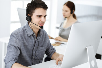 Casual dressed young man using headset and computer while talking with customers online. Group of...