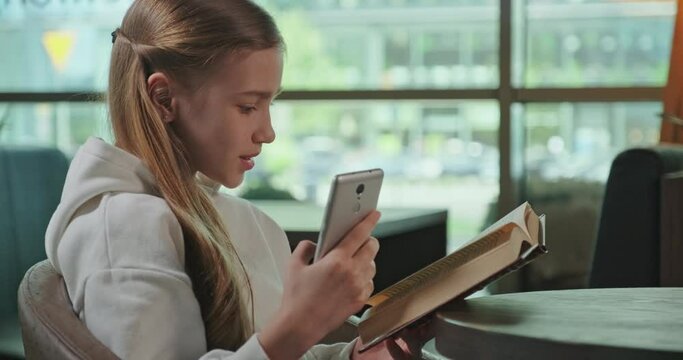 Profile portrait blond preteen girl 10s at the same time reading book and using cell phone inside cafe