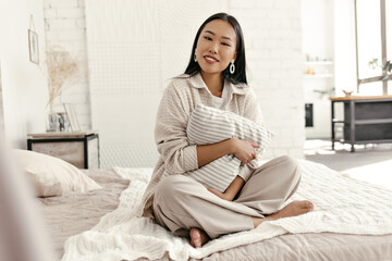 Obraz premium Attractive brunette lady in beige cardigan and pants smiles, looks into camera and hugs pillow in bedroom. Young Asian woman in stylish outfit sits on soft bed.