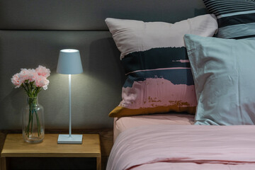 Modern bedside table bed lamp in cozy bedroom with pink carnations flowers in a glass bottle and modern fabric pillows interior beautiful living room decoration home architecture.