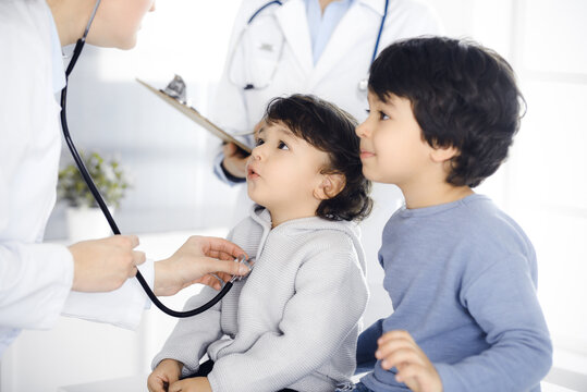 Woman-doctor examining a child patient by stethoscope. Cute arab toddler and his brother at physician appointment. Medicine concept