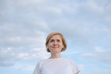 A young Caucasian girl in a white T-shirt and headphones stands against the background of a blue sky with clouds. The concept of listening to music, podcasts. Minimalism, copy space.