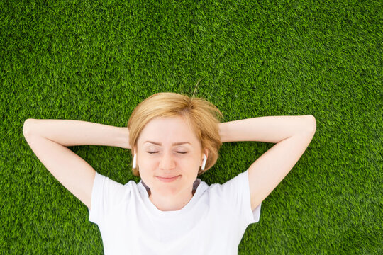 A young Caucasian girl in a white T-shirt and headphones lies on an artificial lawn with closed eyes. The concept of listening to music, podcasts. Minimalism, copy space, top view.