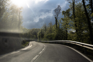 Empty concrete road between green trees under dark clouds and sun, highway before the storm, Catalonia, Spain