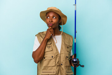 Young african american fisherwoman holding rod isolated on blue background looking sideways with doubtful and skeptical expression.