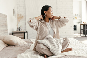 Happy tanned woman in beige jacket and pants sits on soft bed with striped pillow. Cheerful lady in...