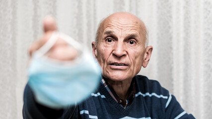 Older male patient holds facemask in hand and shows it with resentment.
