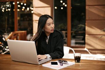 Long-haired brunette woman in black stylish jacket sits at wooden desk and works with laptop. Asian lady poses in street cafe with coffee cup and phone.