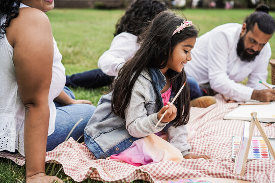 Happy Indian Family Having Fun Doing Picnic While Painting With Children Outdoor At City Park - Main Focus On Girl Face