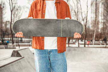 Skater boy in a white T-shirt, brown shirt and blue jeans holds a skateboard in front of him in a...