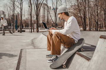 A guy works on his laptop during a break in skiing at the skatepark, which is located in the middle of the spring / autumn city, communicates on social networks and establishes social contacts