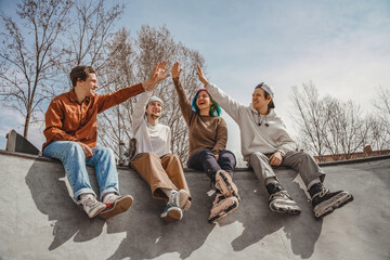 Four rollerbladers and skateboarders, three boys and one girl, sit on a ramp in a city skatepark and give five to each other, cheering up after a fun workout