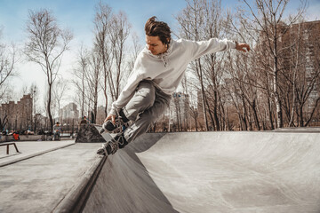 A roller skater in a white sweatshirt rides on the edge of a ramp holding his right foot with his right hand in a gray skatepark in the background a metropolis with an autumn / spring park