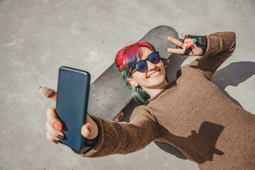 Happy teen girl with colorful hair takes a selfie photo on mobile phone smiling while lying on a skateboard and shows a hand gesture of peace