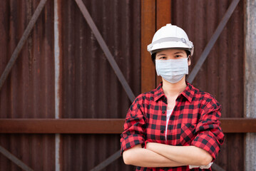 Medium shot portrait with copy space of Asian woman, female worker wearing white safety hard hat, red lumberjack shirt and a mask, standing near rusty metal wall at construction site, crossing arms. 