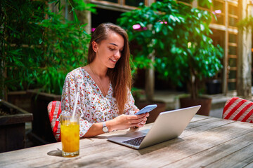 Fototapeta premium Happy cheerful smiling beautiful cute joyful young millennial girl using laptop and phone for remote work, chatting, browsing online and social networking at green cafe