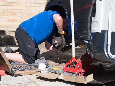 A mechanic has jacked up a motorhome recreational vehicle and removed a wheel and peers under the wheel 
 arch checking the brakes.Trolley jack, socket set are visible