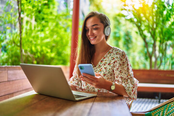 Happy cheerful smiling beautiful cute joyful young millennial freelancer girl wearing wireless headphones using laptop and phone for remote work and online browsing at green cafe