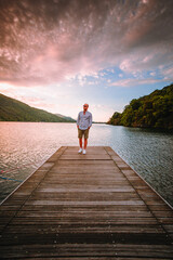 Mergozzo, Verbania / Italy - June 2021: Young man walking on the wooden jetty of Lake Mergozzo at dawn with colorful clouds in the background