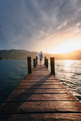 Orta San Giulio / Italy - June 2021: Young man at the bottom of a wooden jetty on Lake Orta as he watches the sun set behind the mountains at sunset