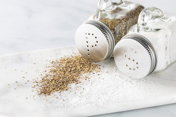 Close up view of salt and pepper shakers tipped over with salt and pepper poured out onto a white marble platter.