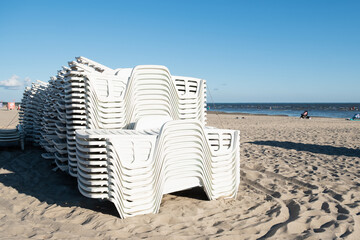 Beach chair renting business closed during summer. White plastic beach chairs stacked together on a sandy beach. Sun loungers rental closed.