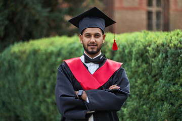 Portrait of indian handsome male graduate in graduation robe with crossed arms and blured background