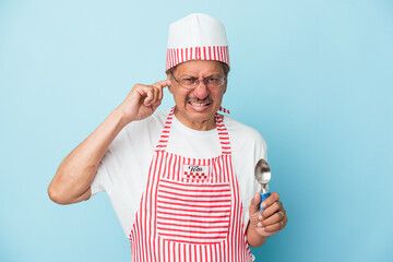 Senior indian ice cream man holding a scoop isolated on blue background covering ears with hands.