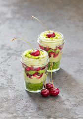 Summer dessert in a glass, trifle of layers of spinach sponge cake, cottage cheese cream with green tea matcha and fresh cherries, on a dark gray background