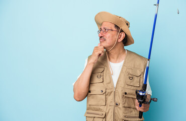 Senior indian fisherman holding rod isolated on blue background looking sideways with doubtful and skeptical expression.