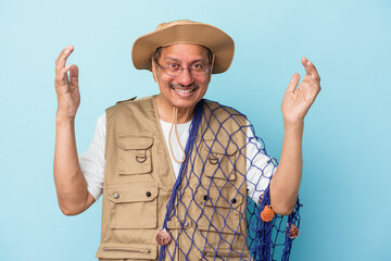 Senior indian fisherman holding net isolated on blue background receiving a pleasant surprise, excited and raising hands.