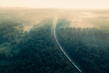 Train in summer morning forest at fog sunrise. Aerial view of moving freight train in forest....