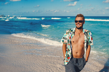 Attractive young man wearing hawaiian shirt at the sea or the ocean background. Travel vacation holiday. Man walking at the sea, enjoy tropical season - obrazy, fototapety, plakaty