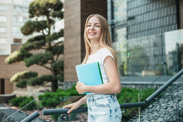 Caucasian blonde student girl holds folders, notebooks in hands and smiles on the background of the university building. Copy space