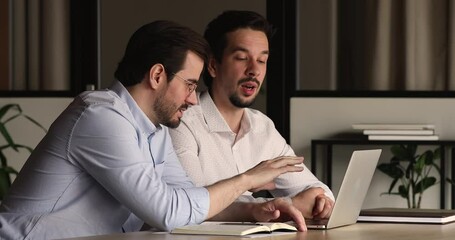 Male colleagues sitting at desk in front of laptop working together on common online project, discuss detail, learn e corporate program. Mentor help to apprentice with new business application concept