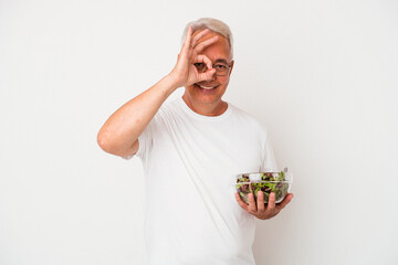 Senior american man eating salad isolated on white background excited keeping ok gesture on eye.