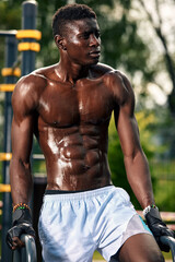 Muscular young man exercising at the sports field. African man looking to the side while doing horizontal bar exercises. Shirtless male model exercising outdoors.