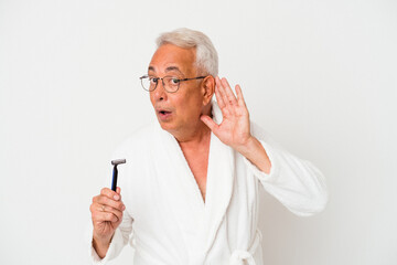 Senior american man wearing bathrobe holding razor blade isolated on white background trying to listening a gossip.