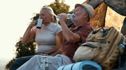 Aged camper couple takes a break from active mountain exploration,  drinking water in front of the tent and enjoying the view from the top