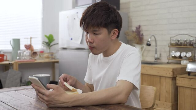 young Asian man holding a mobile phone at the dining table is reading online news with concentration while eating breakfast in the morning.