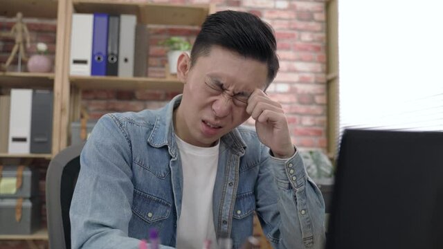 asian man sitting behind a laptop is touching and blinking his eyes while suffering eye irritation at work in the office.