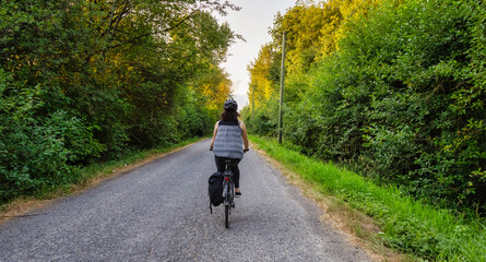 Fototapeta premium Adventurous White Cacasusian Woman riding a bicycle on a road. Sunny Summer Sunset Art Render. Barnston Island, Vancouver, British Columbia, Canada. Adventure Journey Concept