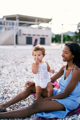 Fototapeta na wymiar Little girl gnaws pebbles while standing next to sitting mother on the beach