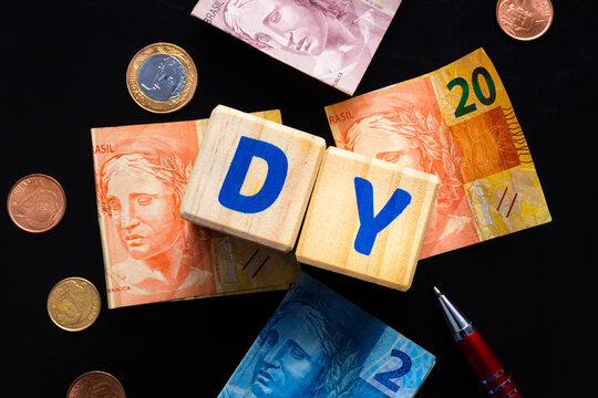 Palavra DY de Dividend Yeld, escrita em cubos de madeira em mesa de cor escura com cédulas do Real brasileiro ( BRL ) e algumas moedas. Conceito para Economia, Mercado de ações e bolsa de valores.