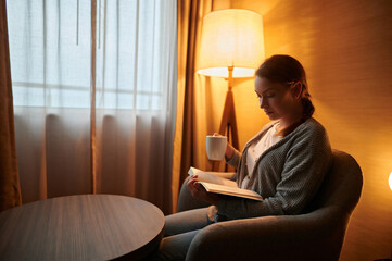 Cozy evening at home.A young woman is sitting in a chair and reading a book. She has a cup of tea...