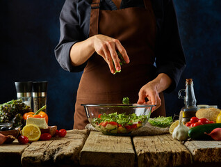 Greek salad preparation process. In a glass bowl, all the ingredients for the salad are cut into pieces. The cook mixes them up. All the ingredients for the salad are next to it. Dark blue background.