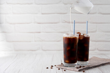 Iced coffee in a tall glass with cream on a white wood background. Cold tasty summer refreshment beverage concept. Selective focus, copyspace.
