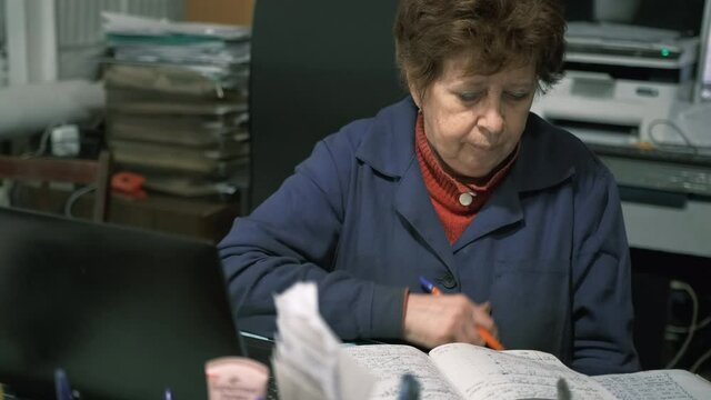 An elderly female engineer sits at a desk heaped with papers and takes notes in a workbook. Vintage accountant searches for information in his records in production