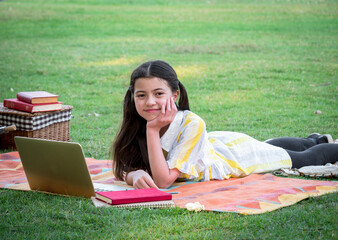 Smart Caucasian girl plays with a laptop, lying on the grass at the park with a smiling face, kid, and technology concept..