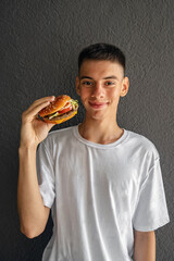 young man of teenage age smiles holding an appetizing burger on the background of gray concrete wall on the street, the concept of the negative impact of fast food on the younger generation